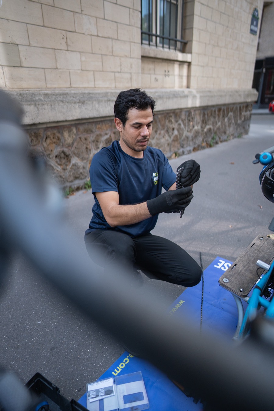 Réparateur vélo Nicolas examinant pièce transmission gants noirs cargo bleu Toulouse
