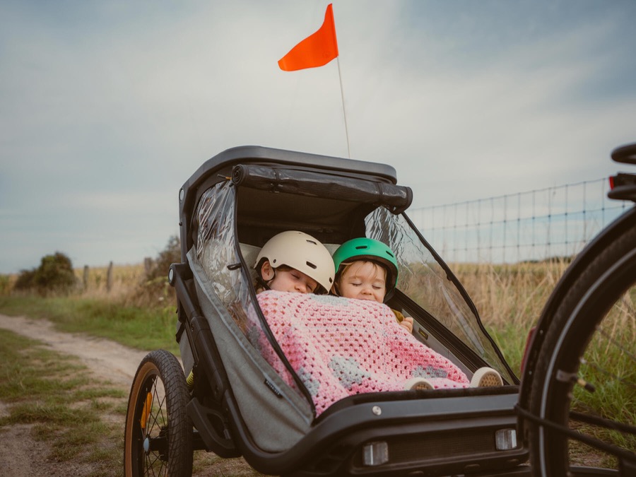 Deux enfants casqués bien protégés dans la cabine d'une remorque vélo enfant, fanion orange déployé pour la visibilité