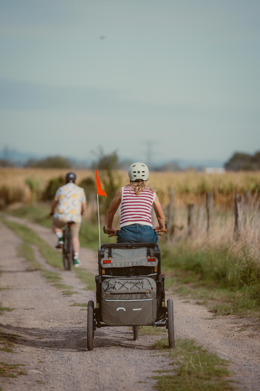 Vue arrière d'une cycliste tractant une remorque vélo enfant sur un chemin de campagne, son compagnon devant